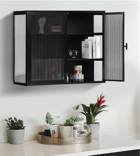 Black metal wall cabinet with fluted glass doors open showing two shelves with books and decor, mounted on white wall